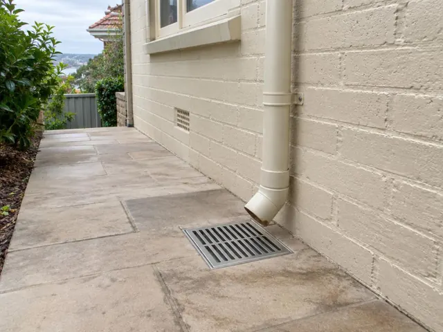 Outdoor drain grate beside a house wall and downpipe on paved courtyard in Dover Heights