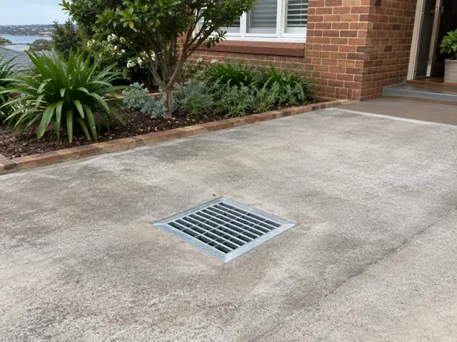 Concrete driveway with a seated stormwater grate beside a brick house and garden in Dover Heights